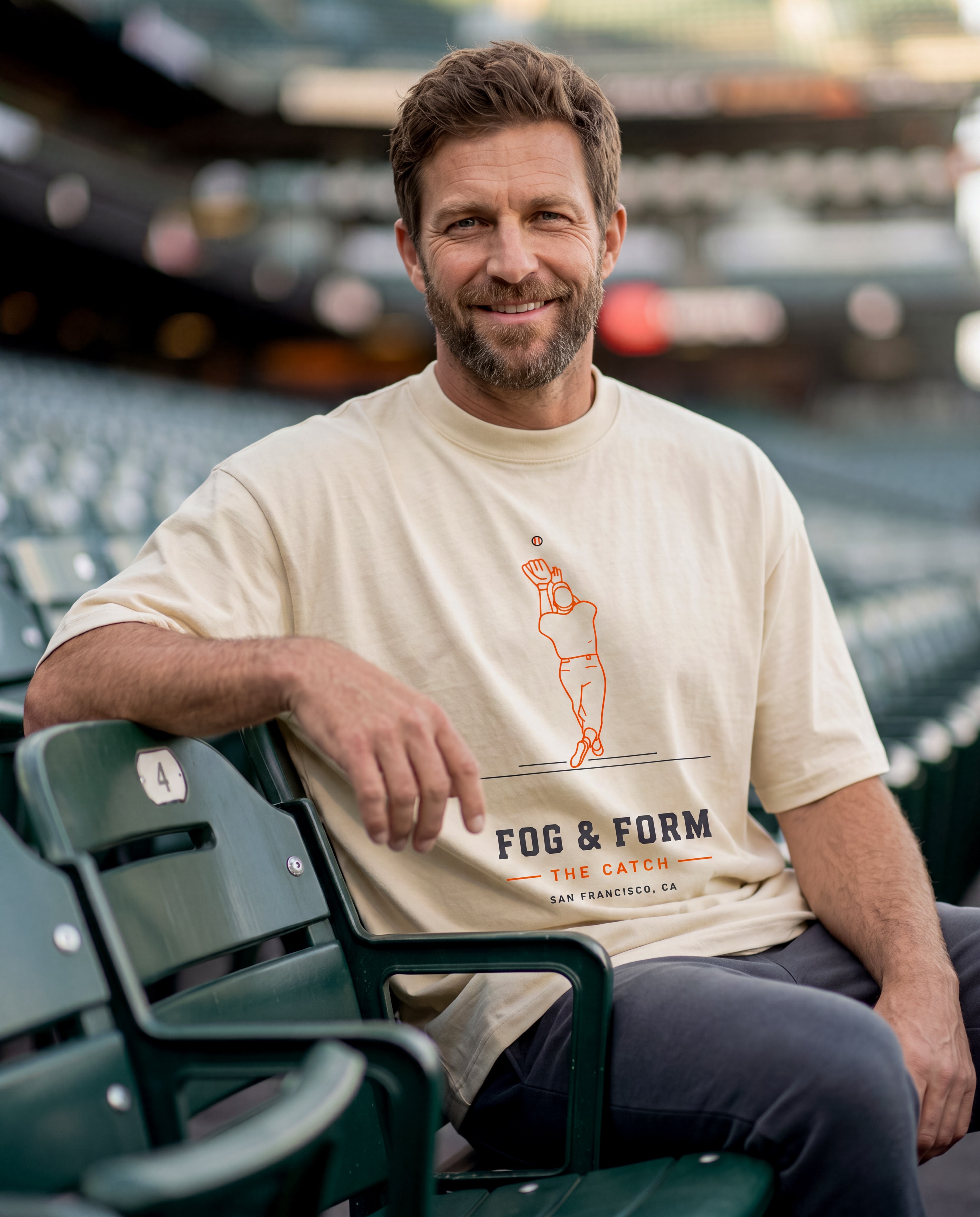 Man sitting on green seats at baseball stadium wearing beige Fog & Form shirt of baseball player catch.