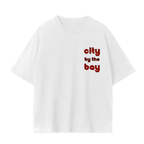 A white short-sleeve t-shirt with the text 'city by the bay' printed on the left chest in a red and black retro font.