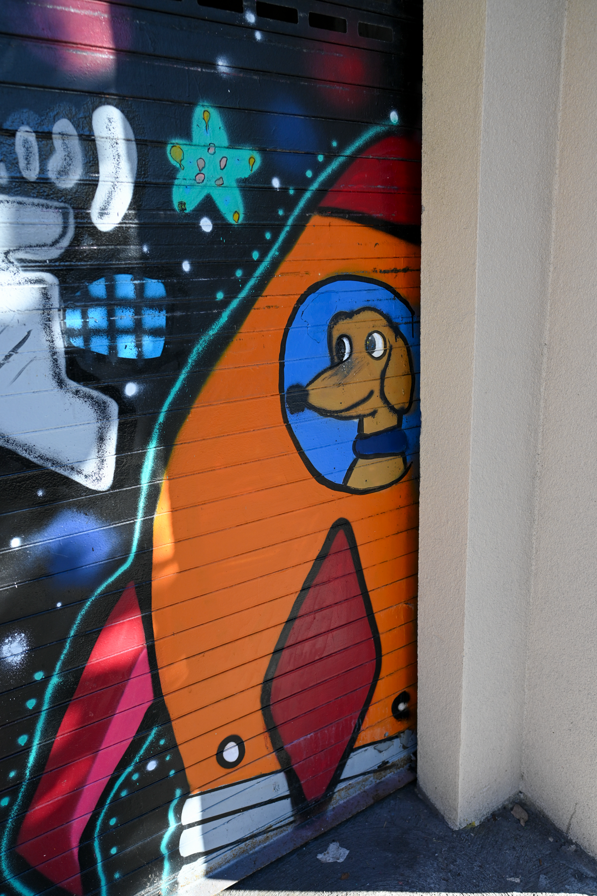 A close-up of a colorful graffiti mural painted on a metal roller door next to a beige wall. The mural features a large orange rocket ship with a cartoon dog character in a blue circular window, alongside other stylized graffiti elements.