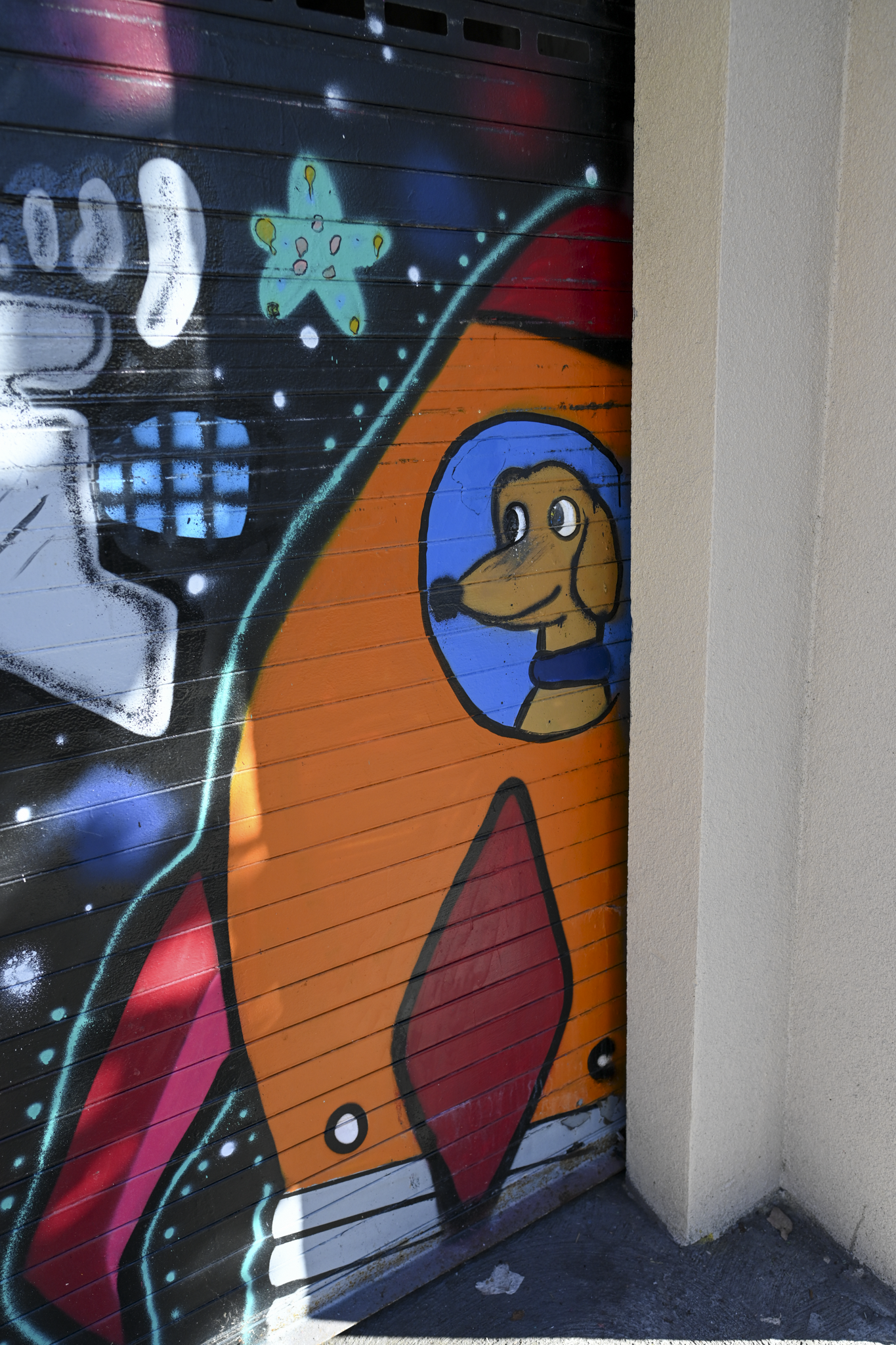 A close-up of a colorful graffiti mural painted on a metal roller door next to a beige wall. The mural features a large orange rocket ship with a cartoon dog character in a blue circular window, alongside other stylized graffiti elements.