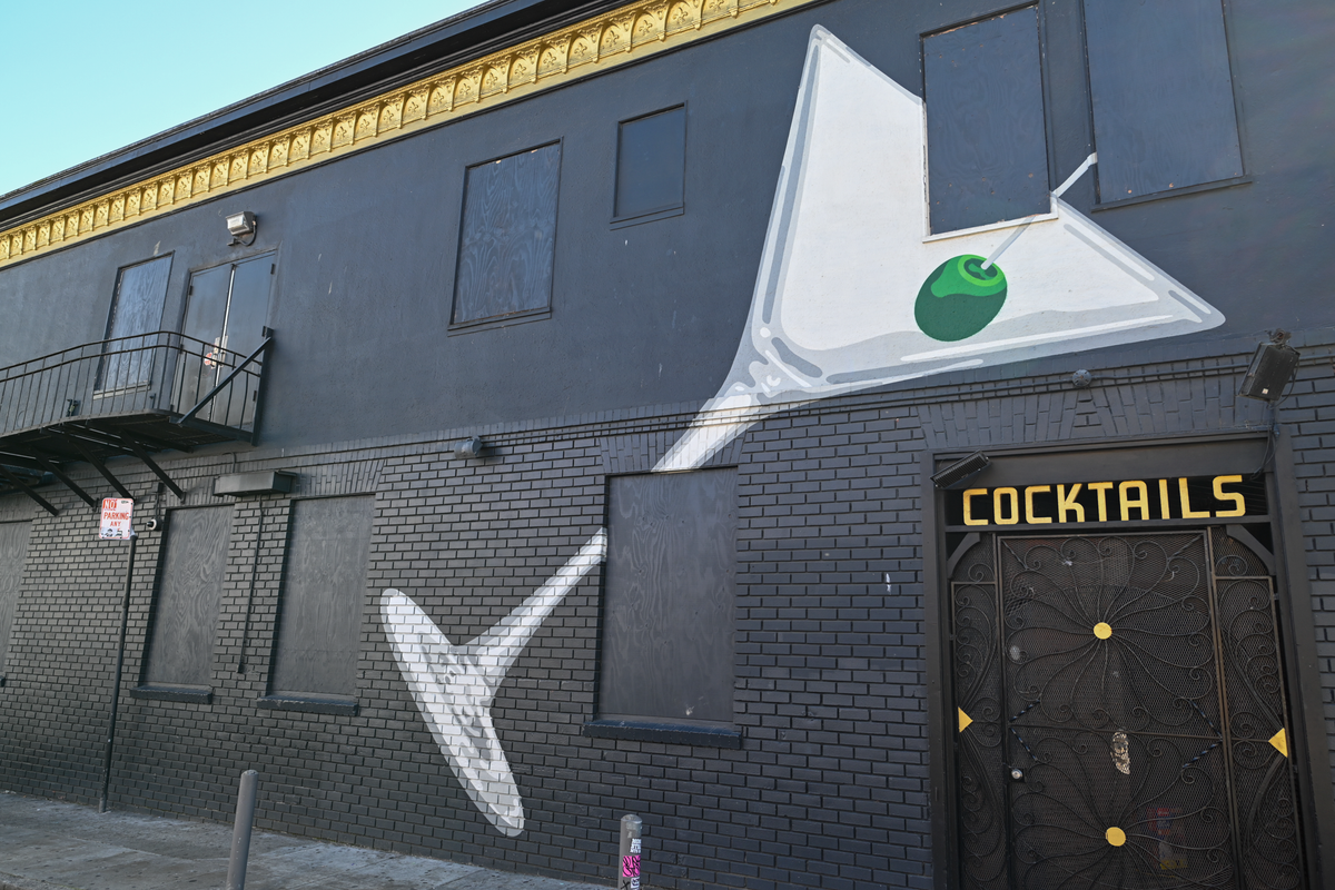 A black brick building facade with a large painted martini glass mural on the wall, featuring a green olive on a toothpick. The word "COCKTAILS" appears in gold letters above a dark door with gold accents.