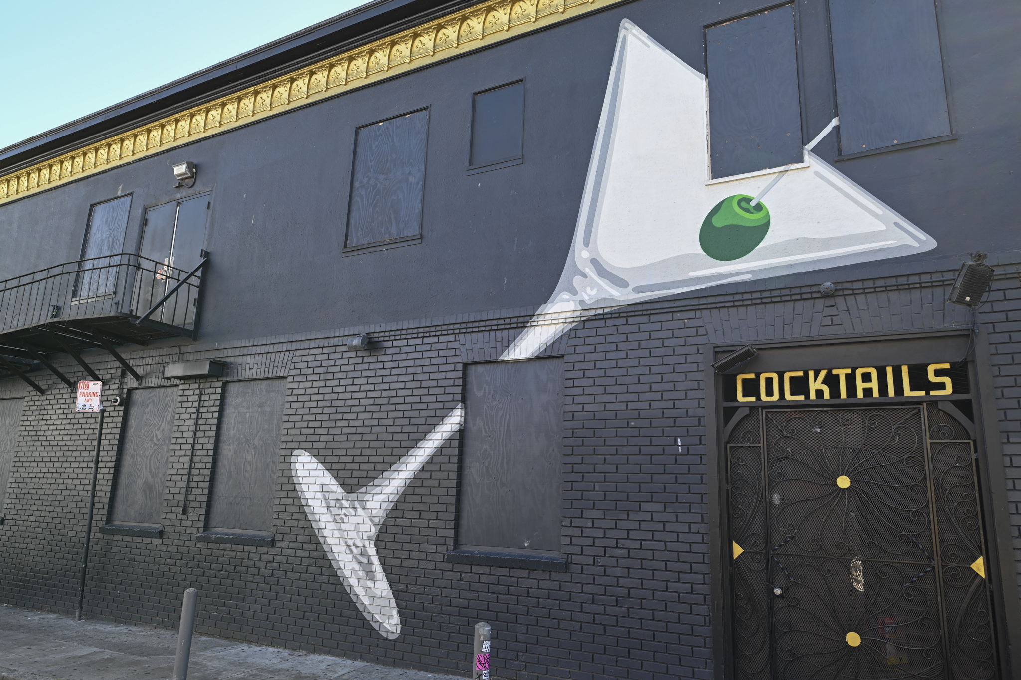 A black brick building facade with a large painted martini glass mural on the wall, featuring a green olive on a toothpick. The word "COCKTAILS" appears in gold letters above a dark door with gold accents.