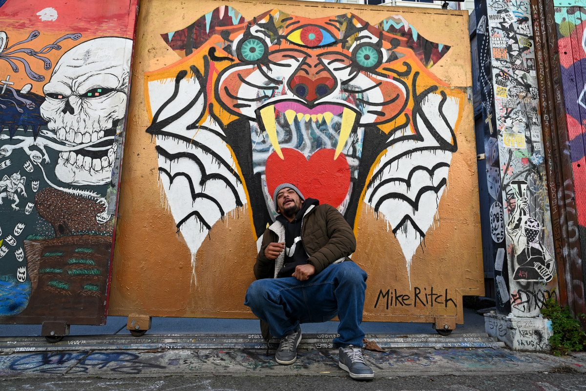 A man wearing a beanie, jacket, and jeans sits in front of a large graffiti mural featuring a stylized tiger face with a red heart on its chest. The mural is signed 'Mike Ritch' and is photographed outdoors between other colorful street art panels.