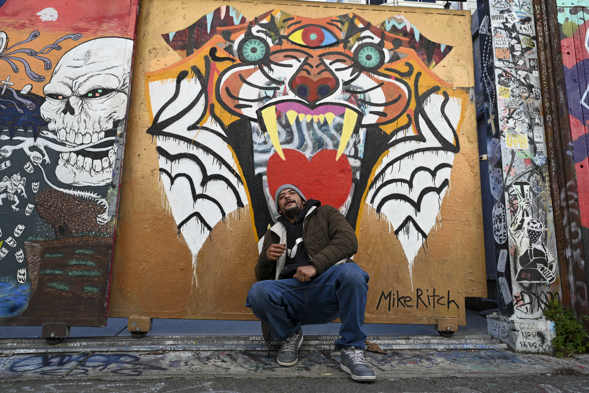 A man wearing a beanie, jacket, and jeans sits in front of a large graffiti mural featuring a stylized tiger face with a red heart on its chest. The mural is signed 'Mike Ritch' and is photographed outdoors between other colorful street art panels.