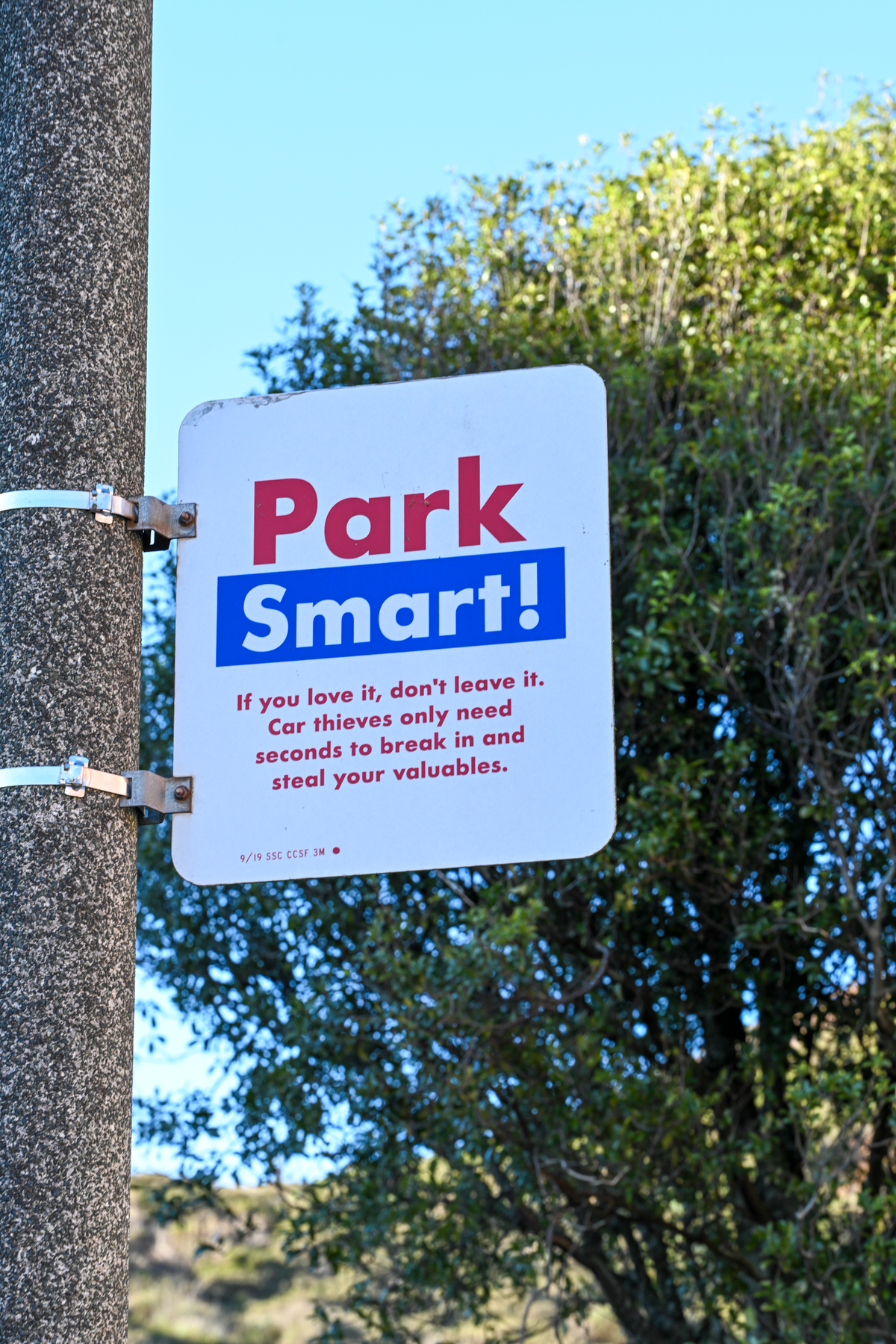 A white rectangular sign reading 'Park Smart!' in red and blue text, mounted on a gray concrete pole outdoors with trees and blue sky in the background.