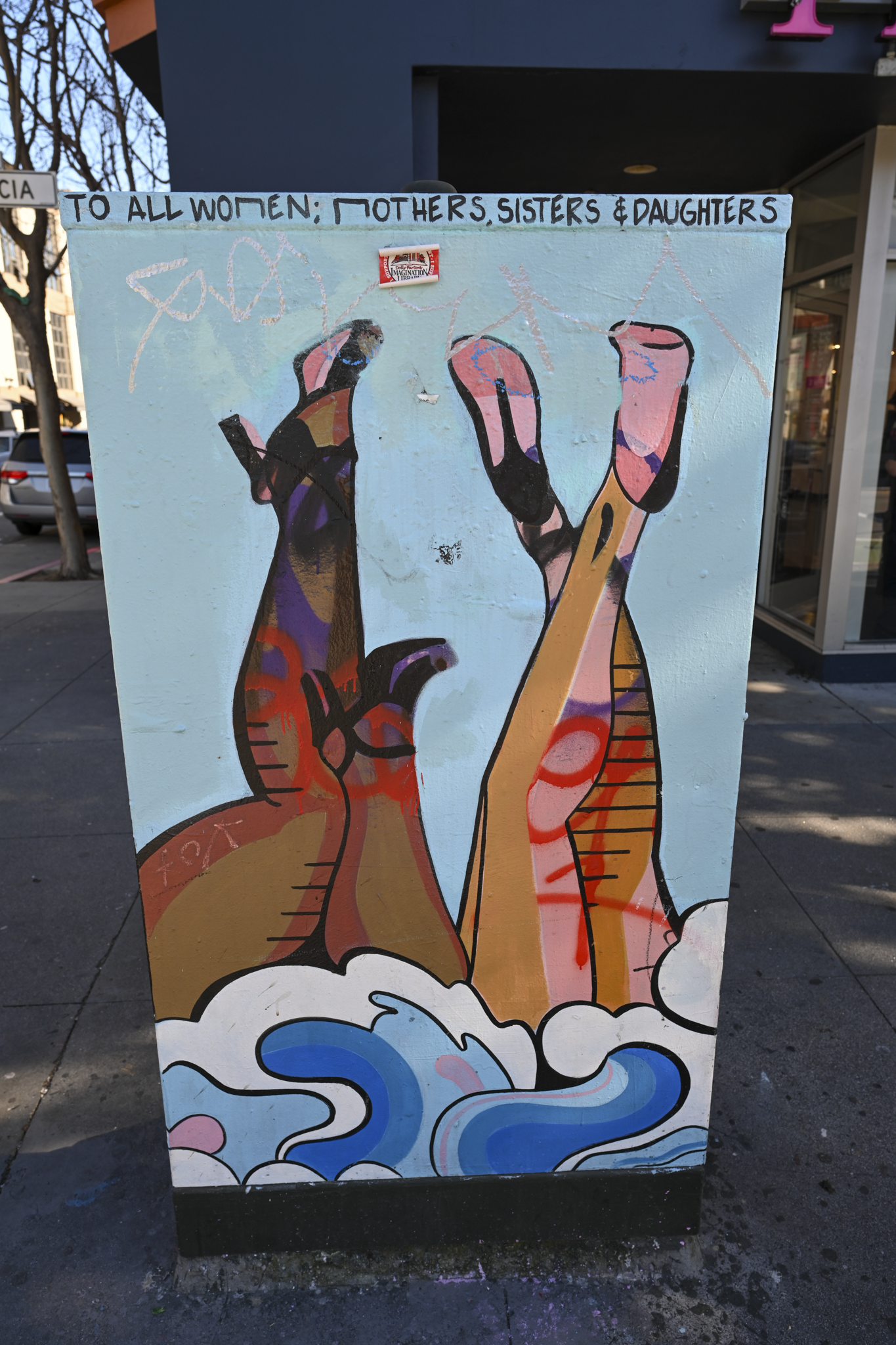 A street-art painted utility box on a city sidewalk, featuring stylized legs in pink, orange, and purple above blue waves, with the text 'TO ALL WOMEN; MOTHERS, SISTERS & DAUGHTERS' at the top.