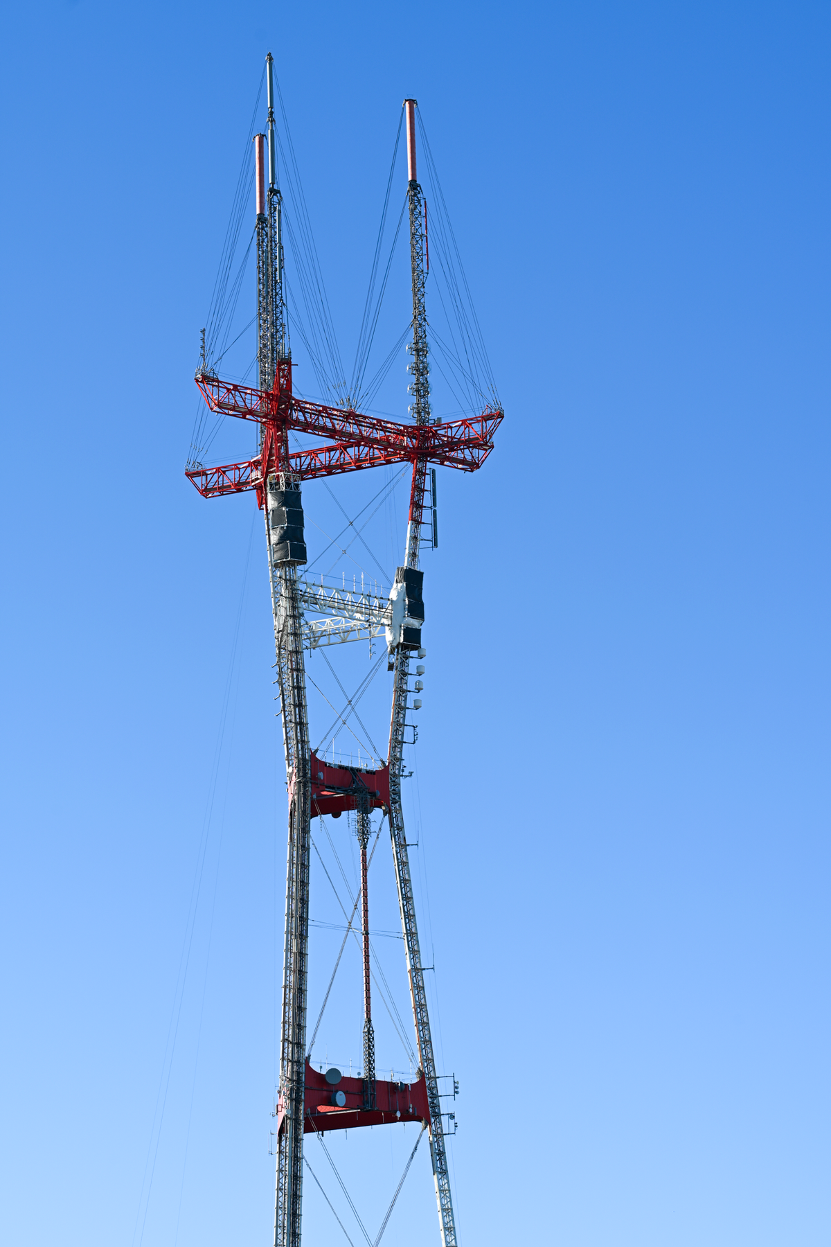 A tall metal lattice Sutro  tower with red structural crossbeams and cables, photographed against a clear blue sky.