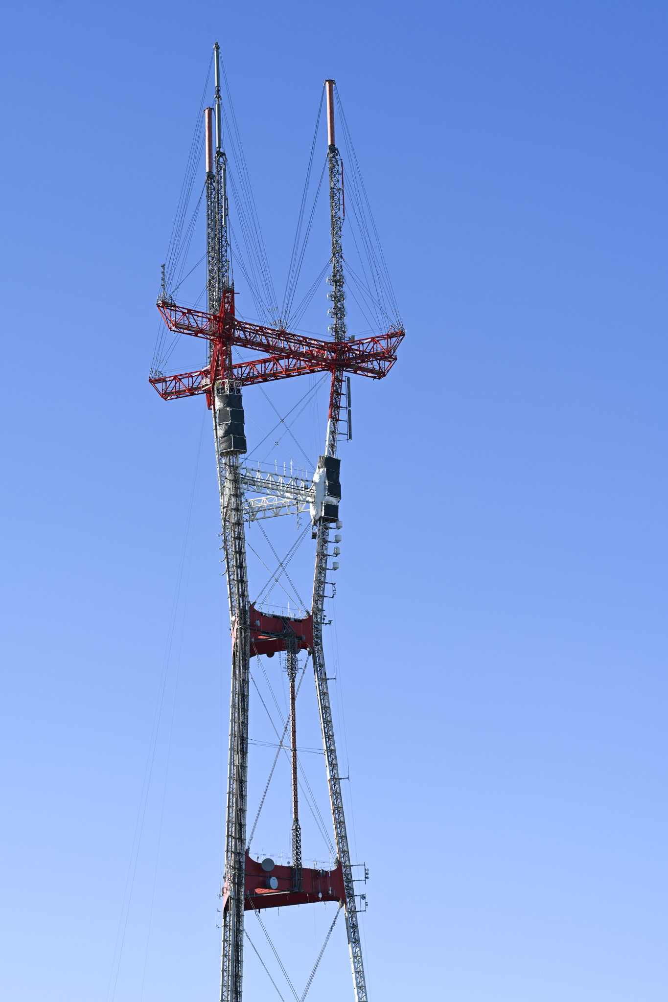A tall metal lattice Sutro  tower with red structural crossbeams and cables, photographed against a clear blue sky.
