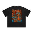 A black short-sleeve t-shirt with a large colorful graphic on the back. The graphic features the words 'BE GRATEFUL' in orange, a skull with a floral crown, a skeleton, a house, and blue waves for SF music.