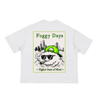 A white short-sleeve t-shirt with the graphic of Karl the Fog wearing a green cap and sunglasses, smoking, with a city skyline and Golden Gate Bridge in the background.